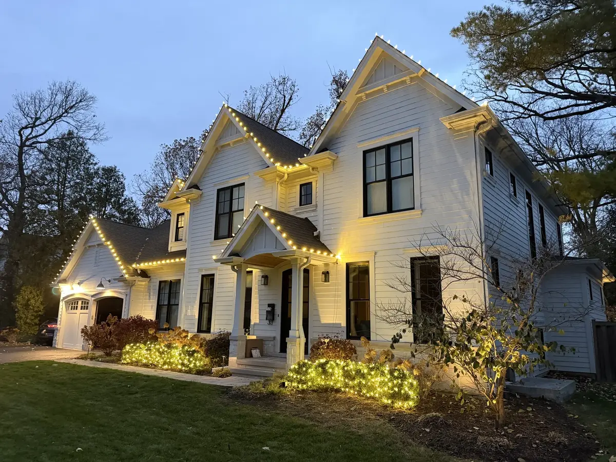 White modern farmhouse with warm roofline lights and lit bushes