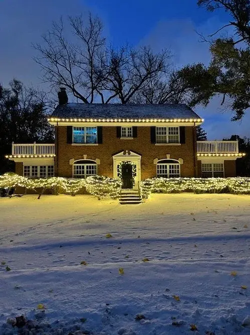 Snow-covered colonial with roofline lights and lit hedges